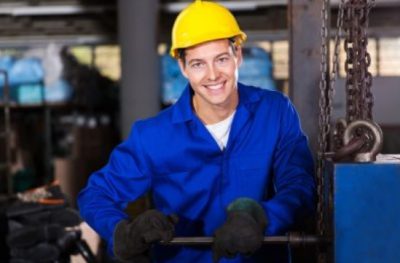 A man in a blue hard hat is smiling in a factory, working as a diesel mechanic.